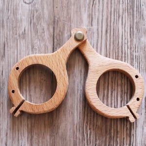 Puede incluir: Un par de monturas de gafas de madera, unidas en el puente con un remache metálico. Las monturas son de color marrón claro, con aberturas circulares para las lentes y una ranura en la parte inferior de cada montura. La veta de la madera es visible.