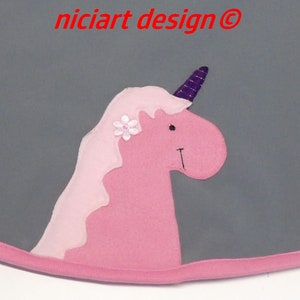 May include: A pink unicorn applique with a purple horn and a white flower on a gray background. The unicorn is a whimsical design with a friendly face and a wavy mane.