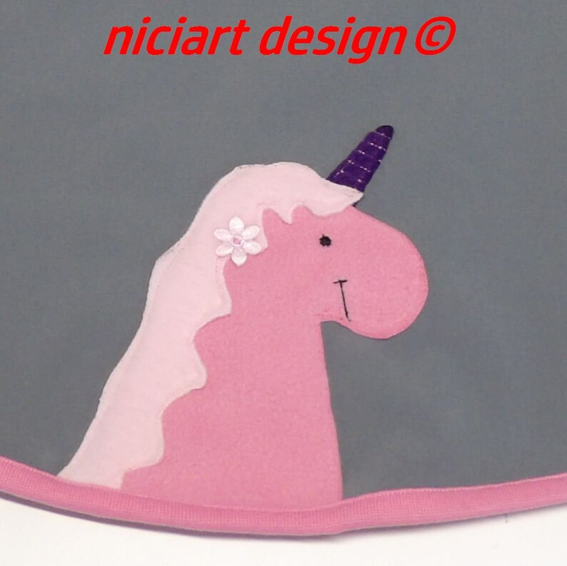 May include: A pink unicorn applique with a purple horn and a white flower on a gray background. The unicorn is a whimsical design with a friendly face and a wavy mane.