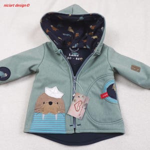 Può includere: Una giacca verde con cappuccio con un design di tricheco blu e bianco. La giacca ha una cerniera e una tasca sul davanti. Il testo "LASS 80 - 86" è stampato all'interno del cappuccio.