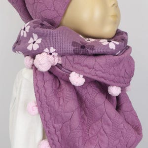 Puede incluir: Un conjunto de gorro y bufanda de punto morado con estampado floral y pompones rosas.