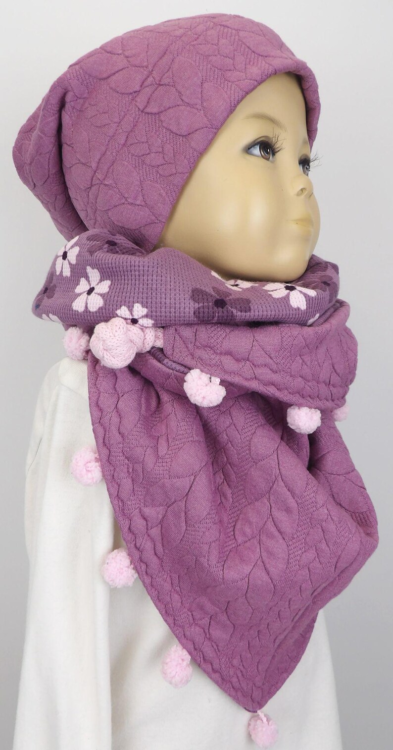 Puede incluir: Un conjunto de gorro y bufanda de punto morado con estampado floral y pompones rosas.