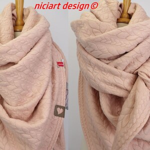 Op de afbeelding: Een lichtroze gebreide sjaal met een hartjespatroon. De sjaal is gevouwen en wordt getoond op een mannequin.