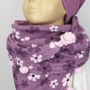 Puede incluir: Una bufanda de punto morado con un patr&oacute;n floral y pompones rosas. La bufanda la lleva un ni&ntilde;o que lleva un gorro de punto morado con un logotipo en forma de coraz&oacute;n.