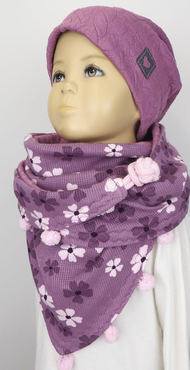 Puede incluir: Una bufanda de punto morado con un patr&oacute;n floral y pompones rosas. La bufanda la lleva un ni&ntilde;o que lleva un gorro de punto morado con un logotipo en forma de coraz&oacute;n.