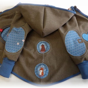 Può includere: Una giacca con cappuccio marrone con dettagli blu. La giacca ha un cuore blu, due cerchi blu con pois bianchi e due cerchi blu con animali ricamati.