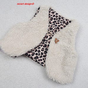 Könnte beinhalten: Eine cremefarbene Kunstfellweste mit Leopardenmusterfutter. Die Weste hat einen kleinen, braunen, herzförmigen Knopfverschluss. Der Text "niciart design ©" befindet sich in der oberen linken Ecke.