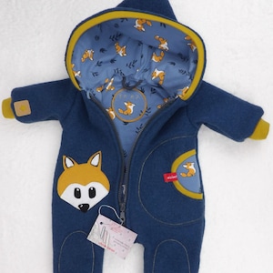 Op de afbeelding: Een blauwe baby romper met een vos ontwerp. De romper heeft een capuchon met een blauwe en witte vos print voering. De romper heeft een ritssluiting aan de voorkant en een gele bies rond de capuchon en zakken.