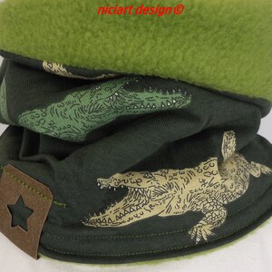 Pode incluir: Cachecol verde com forro de lã e estampa de crocodilos. O cachecol tem uma etiqueta de couro marrom com uma estrela.