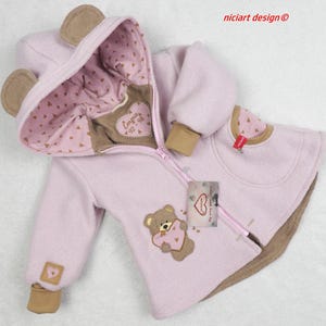 Könnte beinhalten: Hellrosa Babyjacke mit Kapuze und Bärenohren. Die Jacke hat einen braunen Teddybär-Applikation, der ein Herz hält. Das Innenfutter der Kapuze und die Manschetten sind mit kleinen braunen Herzen gemustert. Die Jacke hat einen Reißverschluss.