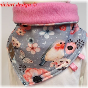 Peut inclure: Un tour de cou en tissu gris et rose avec un imprimé floral et un oiseau. Le tour de cou est doublé de polaire rose et comporte une petite pièce en tissu marron avec un cœur. Le texte "niciart design ©" est visible dans le coin supérieur gauche de l'image.