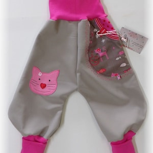 Può includere: Pantaloni da bambino grigi con bordino rosa e un'applicazione di gatto rosa. I pantaloni hanno una tasca con una stampa di gatto e pesce.