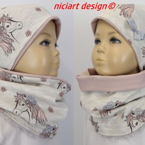 Könnte beinhalten: Eine graue und rosafarbene Strickmütze und ein passender Infinity-Schal mit einem Einhorn-Druck. Die Mütze hat eine rosafarbene Schleife an der Seite. Der Schal ist reversibel mit einer rosafarbenen Seite und einer grauen Seite.