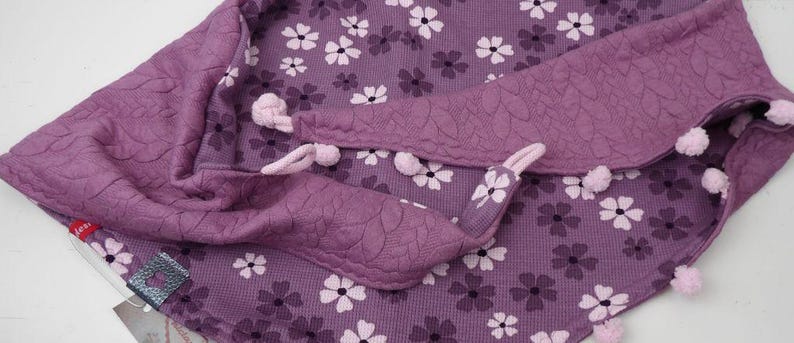 Puede incluir: Bufanda de tela morada con un patr&oacute;n floral y pompones en los bordes.