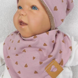 Può includere: Set rosa per neonati composto da cappello e bavaglino con motivo a cuore. Il cappello ha una fodera in pile marrone e il bavaglino presenta un'etichetta in pelle marrone con un cuore ritagliato. Set per neonati.