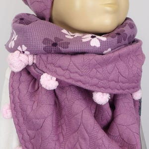 Puede incluir: Un conjunto de gorro y bufanda de punto morado con estampado floral y pompones.