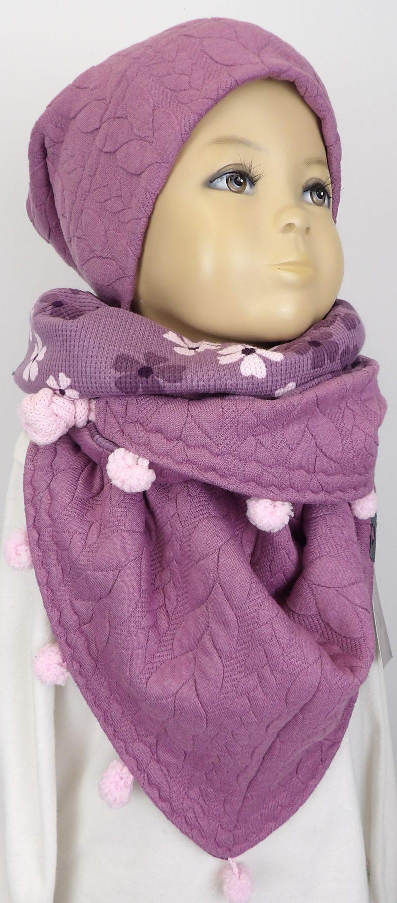 Puede incluir: Un conjunto de gorro y bufanda de punto morado con estampado floral y pompones.