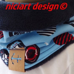 Op de afbeelding: Een blauwe en witte nekwarmer met een rood en wit ankerontwerp. De nekwarmer is gevoerd met een donkerblauwe fleece stof. De nekwarmer heeft een bruin label met een wit anker symbool.