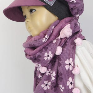 Puede incluir: Un gorro de punto morado con estampado de flores y una bufanda a juego con pompones rosas. El gorro tiene una visera gris y un parche en forma de coraz&oacute;n.