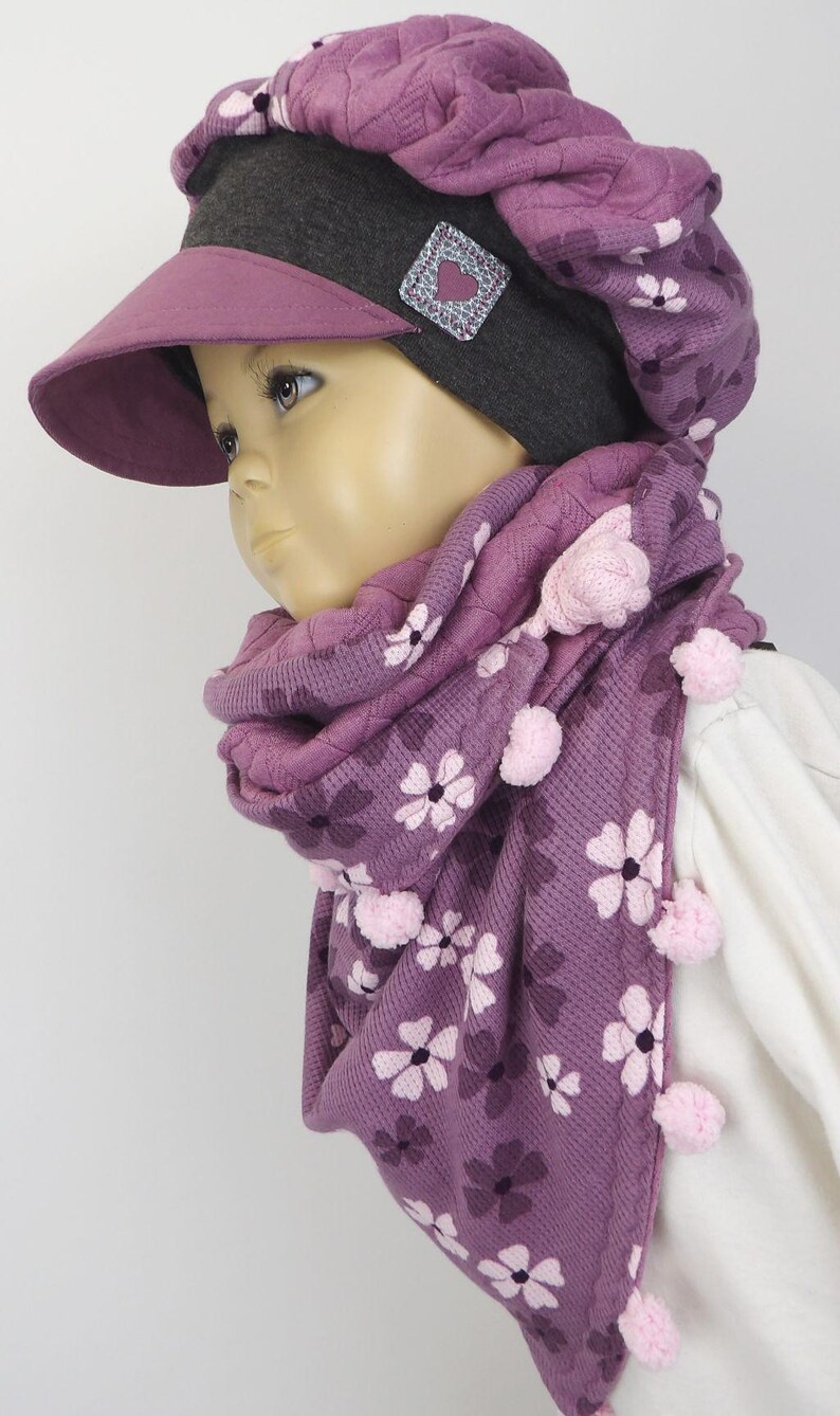 Puede incluir: Un gorro de punto morado con estampado de flores y una bufanda a juego con pompones rosas. El gorro tiene una visera gris y un parche en forma de coraz&oacute;n.