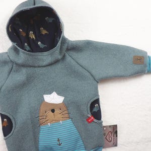 Op de afbeelding: Een tealblauwe hoodie met een zeehond die een matrozenhoed draagt en een blauw-wit gestreept shirt op de voorzak heeft. De capuchon is gevoerd met een donkerblauwe stof met een witte zeehondprint.