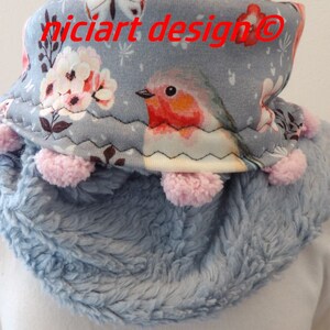 Op de afbeelding: Een lichtblauwe nekwarmer met een pluche textuur en een roze pompon rand. De nekwarmer heeft een bloemen- en vogelprint met de tekst "niciart design@" aan de voorkant.