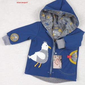 Może przedstawiać: Niebieska kurtka z kapturem z naszywką białego mewą i żółtymi akcentami. Kurtka zapinana na zamek błyskawiczny, z kieszenią z motywem kotwicy. Kaptur podszyty jest tkaniną w szary i biały wzór kotwic.