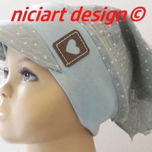 Peut inclure: Un chapeau en tissu bleu clair à pois blancs avec un patch en forme de cœur brun. Le chapeau a une visière et une longue écharpe attachée. Le chapeau est porté par un enfant.