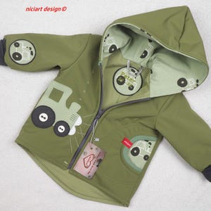 Può includere: Giacca con cappuccio verde oliva con design a trattore. La giacca presenta una chiusura con cerniera, un cappuccio foderato verde chiaro e polsini grigio scuro. Il design del trattore è ripetuto sulla giacca e sul cappuccio.