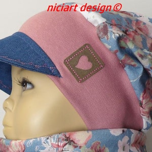 Op de afbeelding: Een roze en blauwe gestreepte beanie met een denim visor. De beanie heeft een bruine leren patch met een roze hart erop. De beanie wordt gedragen door een kind.