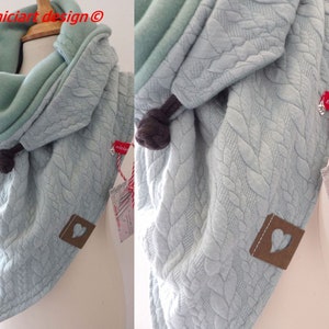May include: A light blue knitted scarf with a fleece lining. The scarf has a cable knit pattern and a heart-shaped tag. The scarf is tied with a brown cord.
