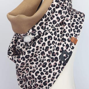 Op de afbeelding: Een capuchonsjaal met een luipaardprint in zwart, bruin en beige. De binnenvoering is effen beige. De sjaal heeft een grijze pompon en een klein leren label. Gepresenteerd op een mannequin. Modieus accessoire.