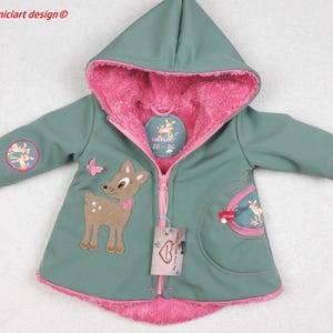 May include: A teal hooded jacket with a pink interior and trim. The jacket features a deer applique, a pink butterfly, and a round patch with a deer illustration. The jacket has a zipper closure and a tag.