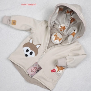 Może przedstawiać: Beżowa kurtka z kapturem z motywem lisa. Kurtka posiada aplikację z lisem z przodu i podszewkę z motywem lisa wewnątrz kaptura. Z zapięciem na zamek błyskawiczny i małą naszywką z napisem "Peter 80/86".