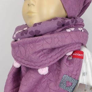 Puede incluir: Un gorro y una bufanda a juego de punto morado con un dise&ntilde;o en forma de coraz&oacute;n. El gorro y la bufanda tienen un patr&oacute;n texturizado y est&aacute;n decorados con peque&ntilde;as borlas.