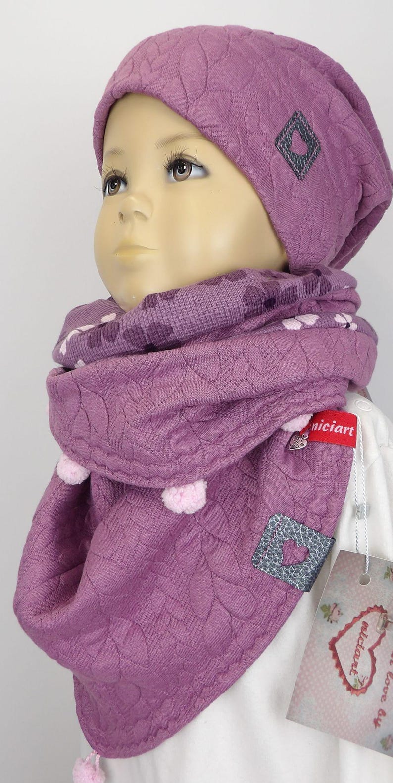 Puede incluir: Un gorro y una bufanda a juego de punto morado con un dise&ntilde;o en forma de coraz&oacute;n. El gorro y la bufanda tienen un patr&oacute;n texturizado y est&aacute;n decorados con peque&ntilde;as borlas.