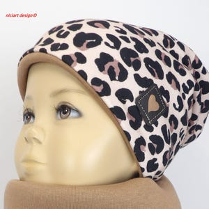 Può includere: Un berretto a maglia con stampa leopardata beige e nera con bordo marrone. Il cappello ha una piccola toppa quadrata in pelle marrone a forma di cuore. Il testo "niclart design" è visibile nell'angolo in alto a sinistra.