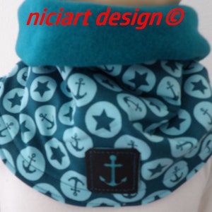 Op de afbeelding: Een teal en marineblauwe infinity sjaal met een nautisch thema. De sjaal heeft een patroon van ankers en sterren in lichtblauwe cirkels. Het bovenste gedeelte is effen teal, en een kleine zwarte patch met een anker is op de voorkant genaaid. De tekst "niciart design" is zichtbaar.