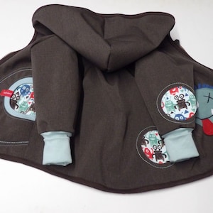 Könnte beinhalten: Eine braune Kapuzenjacke mit blauen und grünen Monster-Patches. Die Jacke hat einen Reißverschluss und eine Kapuze. Die Patches sind an den Ärmeln und am Rücken der Jacke angenäht.