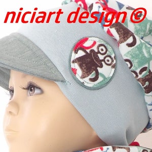 May include: A light blue baby hat with a visor and a brown and green monster patch. The hat has a white and green patterned lining. The text "niciart design@" is visible on the hat.