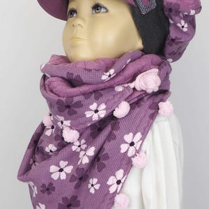 Puede incluir: Una bufanda con estampado morado y rosa con pompones rosas. La bufanda la lleva un ni&ntilde;o que lleva un gorro morado y gris con visera.