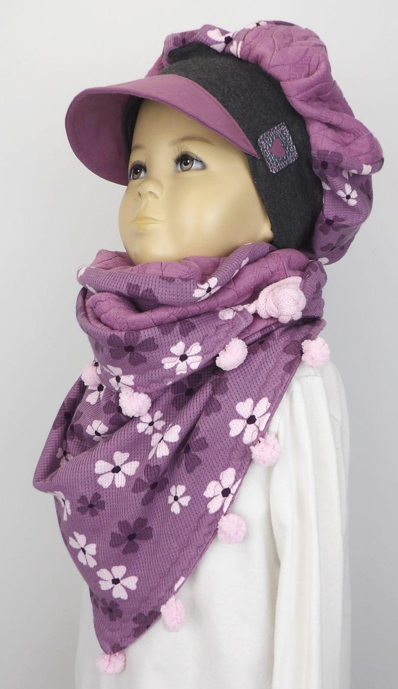 Puede incluir: Una bufanda con estampado morado y rosa con pompones rosas. La bufanda la lleva un ni&ntilde;o que lleva un gorro morado y gris con visera.