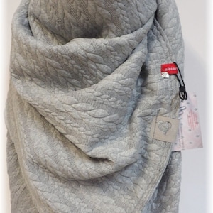 May include: Gray knitted scarf with a cable knit pattern. The scarf is folded and draped over a mannequin. The scarf has a small tag with the word "niciar" on it.