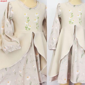 Op de afbeelding: Een beige jurk met lange mouwen en een madeliefjesprint op de mouwen en rok, met een effen beige lijfje voorzien van geborduurde witte en gele madeliefjes. De rok heeft een gelaagd ontwerp met een split aan de voorzijde en elastische boorden.