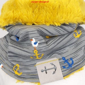 Può includere: Un scaldacollo grigio e giallo con un bordo giallo soffice. Il tessuto ha un tema nautico con ancore e gabbiani. Il testo "nicicart design ©" è visibile nella parte superiore centrale dell'immagine.