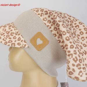 Op de afbeelding: Een beige en bruine luipaardprint hoed met een vizier. De hoed heeft een beige geribbelde band en een kleine, vierkante, bruine patch met een hartuitsparing. De tekst "niciart design ©" staat in de linkerbovenhoek.