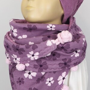 Puede incluir: Bufanda de punto morado con estampado floral y pompones rosas. La bufanda la lleva un ni&ntilde;o que lleva una boina morada con un logotipo cuadrado gris.