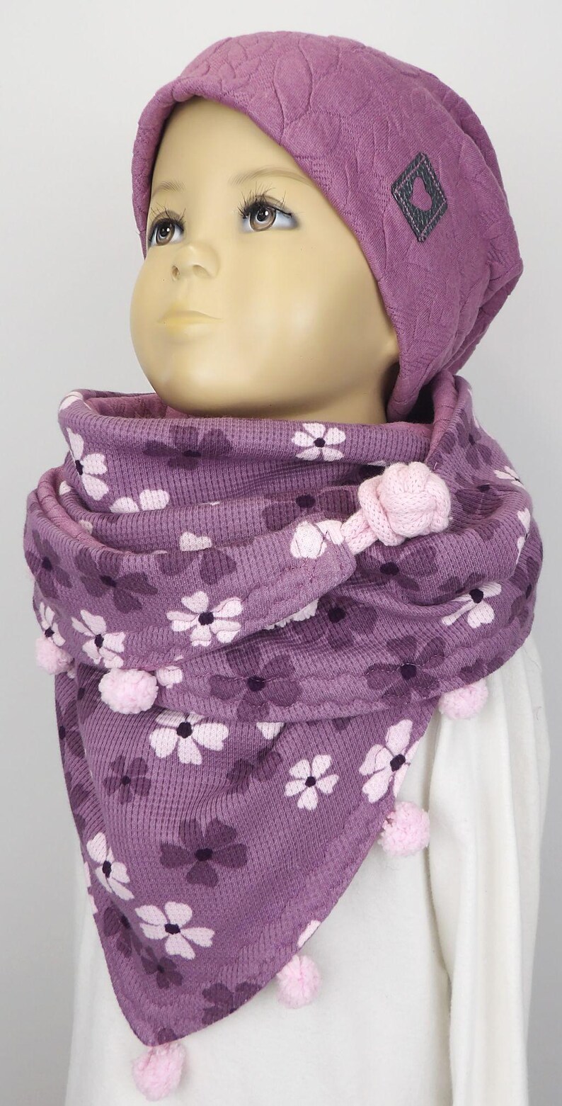Puede incluir: Bufanda de punto morado con estampado floral y pompones rosas. La bufanda la lleva un ni&ntilde;o que lleva una boina morada con un logotipo cuadrado gris.