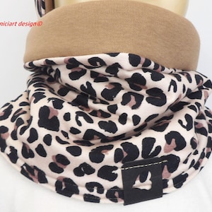 Può includere: Sciarpa tubolare con stampa leopardata beige, nera e marrone. La sciarpa ha una fascia color beige nella parte superiore e un'etichetta in pelle nera con un logo. Il testo "niciart design" è visibile in alto a sinistra.