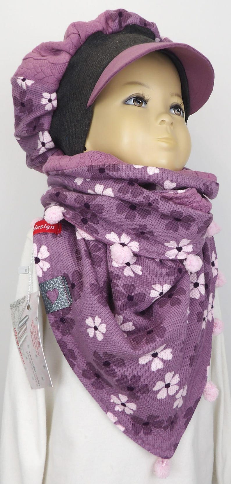 Puede incluir: Una bufanda de punto morada con un patr&oacute;n floral y pompones rosas. La bufanda la lleva un ni&ntilde;o que lleva un gorro morado y gris con ala.